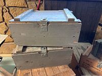 Two stacked vintage military-style wooden trunks painted gray with metal latch and rope handles visible, positioned on a wooden surface against a stone wall background.