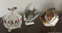 Photo of three glass pieces on table: a vintage covered glass trinket dish with a butterfly handle, a clear crystal fish paperweight, and a tiger art glass figure