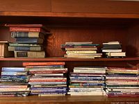 Wide shot showing wooden shelves with multiple stacks of books covering diverse topics including art, painting, woodworking, golf, and history. Books are in various colors and sizes reflecting used condition.