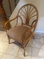 Cane chair with rounded back and armrests placed on tile floor near a wooden post, showing complete chair structure and cushion.