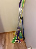 Image shows a group of cleaning tools including two Swiffer mops with green pads and purple handles, two multicolored feather dusters, a yellow mop with a green mop head and white handle, and a black floor squeegee with a blue handle all leaning against a wall on a wood floor.