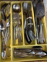 Overview of assorted cutlery pieces including forks, spoons, specialty knife and tongs arranged in a sectioned yellow plastic tray.