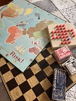 Photo of Risk board game board, wooden peg solitaire game, poker chips tin, and decks of cards on chess/checkers board