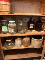 Shelf with antique glass jars including labeled Kraft and Marsel jars, clear glass jars, and amber glass jugs on the top shelf. Bottom shelf has blue-gray ceramic crock, brown stoneware jar, earthenware pot, and glass jar with wooden lid.