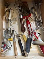 Drawer full of assorted kitchen utensils including rotary hand mixer, whisk, grater, masher, can opener, and slicer.