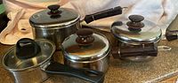 Four pots displayed on a kitchen counter: three Lagostina pots with glass lids and wooden knobs, one ROKitchen saucepan with a glass lid and plastic handle visible. Cloth backdrop.