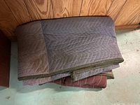 Two folded moving blankets in grey and brown colors, placed against wood panel wall on concrete floor.
