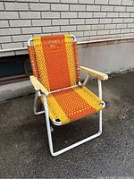 Photo of a single retro folding lawn chair with orange and yellow woven plastic straps, white metal frame, and wooden armrests, placed outdoors near a beige brick wall.