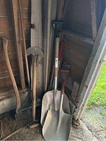 Photo showing several garden tools leaning against a wooden wall inside a shed. Highlights two metal shovels with wooden handles and other tools including rakes and axe.