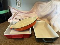 Three stoneware casseroles stacked: rectangular with black handles, square red, oval orange