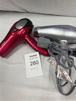 Two hair dryers, one red Conair and one silver John Frieda, both with power cords and tags, laid on a white surface