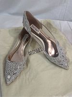 Pair of ivory satin pointy toe flats with crystal embellishments on the toes and vamp area, resting on a Badgley Mischka branded dust bag.