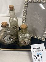 Three glass perfume bottles with cork stoppers and decorative flower accents on a black tray with gold trim, next to a decorative picture frame with pearls and beads on the border.