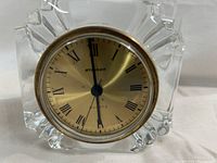 Front view showing gold-tone clock face with black Roman numerals and hands set in clear crystal square body with geometric edge cuts.