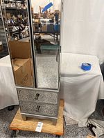 Full view of mirrored jewelry armoire showing upper double cabinet doors with mirrored panels, and two mirrored mosaic drawers below. Surrounding environment visible reflecting in mirror.