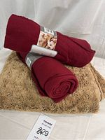 Set of three blankets shown, two red fleece rolled with labels, one tan shaggy plush folded underneath. All blankets stacked together on white background.