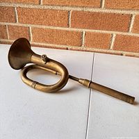 Antique brass car horn resting on a surface with brick wall background, showing full length and curved tube design.