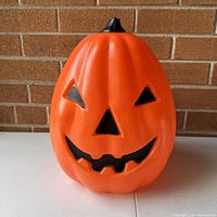 Front view of bright orange plastic pumpkin with black jack-o'-lantern face, smiling with toothy mouth and triangular eyes and nose.