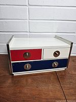 Photo of the closed vintage color block jewelry box showing the red, white, and blue drawers with metallic knobs on a wooden surface against a white brick wall.