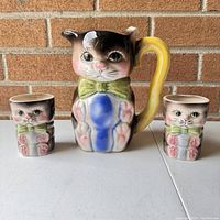 Front view of vintage ceramic anthropomorphic cat pitcher and two kitten cups on white surface against brick wall background