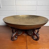 Side view of the brass bowl resting on a carved wooden stand showing the bowl's profile and stand's curved legs and decorative details.