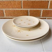Photo of stacked plates, two larger dinner plates and two smaller side plates with gold starburst atomic design on cream base.