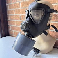 Side view of black vintage army gas mask on mannequin head showing rubber face covering, eye lenses, ventilation cover, and large filter canister.