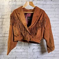 Front view of a brown suede crop jacket with fringe detail hanging on a hanger against a white brick wall.