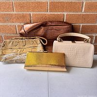 Four vintage purses arranged in a row: bronze faux crocodile pattern bag, snakeskin pattern purse, gold metallic clutch, cream faux crocodile handbag with handle.