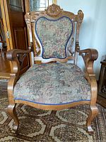 Front view of an antique armchair showing floral tapestry upholstery, carved wooden frame, and metal casters.