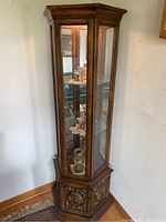 Full view of tall corner curio cabinet showing upper glass display area with glass shelves and mirrored back, sitting on base with carved wood door, in light brown wood finish.