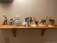 All seven porcelain and ceramic figurines displayed on a wooden shelf against a beige wall, showing their size and detailing.