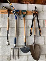 Photo of garden tools including pitchfork, hoe, hand cultivator, hedge shears, and shovel hung on garage wall