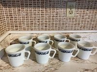 Eight vintage Corelle Old Town Blue Pyrex mugs arranged on a countertop, showing the front side and pattern.
