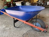 Side view of blue Yard Works wheelbarrow with wooden handles and single black pneumatic wheel on red metal frame.