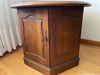 Side view of the round wooden end table showing two paneled cupboard doors with brass handles and decorative carved edges, base with storage.