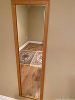 Light wood framed full length mirror hanging on wall showing reflection of floor and rug.