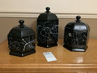 Front view of three black hexagonal ceramic canisters with white paint splashes and black lids with knob handles, arranged side by side.