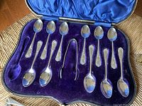 Open leather case showing 5 teaspoons on top row, 6 on bottom, with sugar tongs centered
