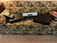 Four electronic devices on floral couch: cassette player, Samsung DVD player, Toshiba DVD/VHS combo player, RCA DVD player.