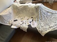 Overview of several table cloths and napkins displayed on black seating furniture, showing beige crocheted cloth and embroidered linens.