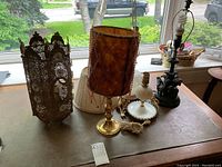Overall view of four lamps including hanging metal lantern, tall shade lamp, milk glass lamp, and elephant base lamp on a wooden surface near window