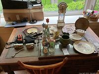 Wide view of the miscellaneous items spread on a table including cameras, plates, decorative items, and stained glass clock near window.