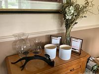 Display of three glass vases of varying sizes, two white ceramic planters, black resin candle holder, two floating picture frames and dry flower arrangement on a wooden side table.