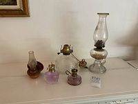 Photo showing entire lot with five oil lamps on surface including finger lamp, miniature lamps, large glass electric lamp, and copper wall oil lamp base.