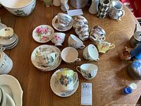 Wide view showing multiple assorted vintage cups and saucers on wooden table with varying floral and scenic designs.