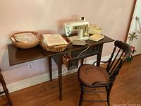 Full view of Singer sewing machine on wooden cabinet, wooden chair beside it, and baskets with sewing supplies on cabinet top.