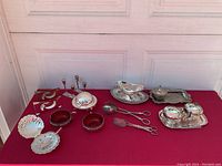 Full view of all silverplate serving pieces arranged on a red tablecloth, includes clam shell butter dish, cream and sugar, candlesticks, serving utensils, napkin holders.