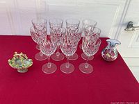Eleven stemmed glasses with embossed diamond and vertical pattern on bowl, a small ceramic flower basket with painted leaves and roses, and a small pottery pitcher with floral decoration on a red cloth surface.