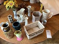 Tabletop display of various antique and vintage ceramic and pottery vases, pitchers, planters, and decorative pieces including artificial flowers and figurines.